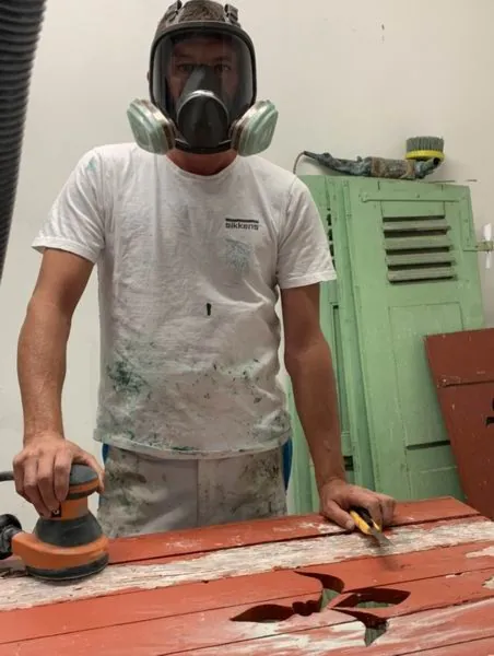 Artisan masqué ponçant un volet bois à la machine dans l’atelier